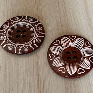 May include: Two brown wooden buttons with white decorative designs. One button has a circular design with concentric circles and the other has a flower design with hearts.