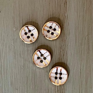 May include: Four round buttons with a white center and gold-colored trim. Each button has six black holes and a decorative gold pattern. The buttons are arranged on a gray wooden surface.