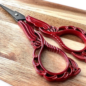 May include: Red decorative scissors with an ornate design. The scissors have a sharp blade and a comfortable handle.
