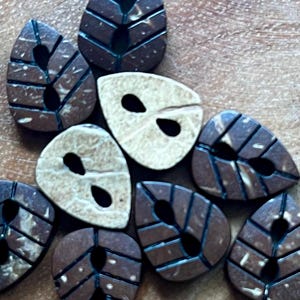 May include: A collection of leaf-shaped buttons in shades of brown and cream. The buttons have a detailed leaf vein design and two holes for sewing. The buttons are made of a natural material and are arranged on a wooden surface.