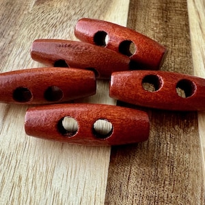 May include: Five wooden toggle buttons in a reddish-brown color. Each button has two holes and a smooth, slightly textured surface.