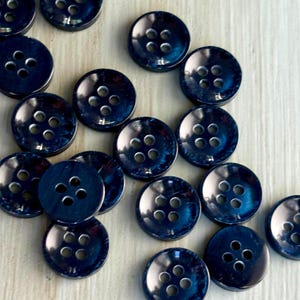 May include: A collection of round, dark blue buttons with a mottled, marbled finish. Each button features a recessed center with four evenly spaced holes for sewing. The buttons have a smooth, glossy surface and are arranged in a scattered pattern.