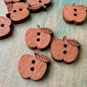 May include: Wooden apple-shaped buttons with two holes and a leaf detail. The buttons are a warm brown color and are scattered on a light blue and green textured surface. These buttons are ideal for sewing and craft projects.