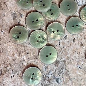 May include: A collection of round, two-hole buttons featuring a shimmering, iridescent light green finish. Each button displays a smooth, polished surface with subtle variations in tone and luster, characteristic of a shell-like material.