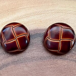 May include: Two round, dark brown buttons with a woven design. The buttons have a glossy finish and are set against a light brown, textured surface. The buttons are likely for clothing or craft projects.
