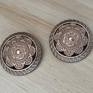 May include: Two antique-style, round, silver-toned earrings with intricate floral and geometric patterns. The earrings have a dark center and a detailed border. The design is symmetrical and ornate, suitable for a vintage or bohemian style.