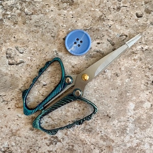 May include: A pair of scissors with iridescent teal and blue handles featuring an ornate, textured design and silver blades, accompanied by a single round, light blue four-hole button with a subtle white swirl pattern on the surface.