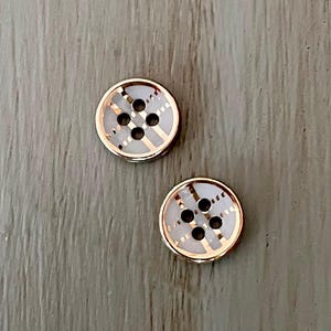 May include: Two round buttons with a gold-colored rim and a white center. The white center has a grid pattern with gold-colored lines and four black holes. The buttons are on a light gray wooden surface.