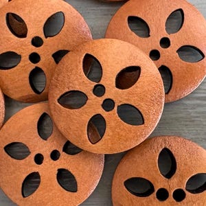 May include: Close-up of several round, orange-brown wooden buttons. Each button has a flower-like design with five oval cutouts and a central hole. The buttons are arranged on a light gray surface.