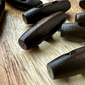 May include: A close-up of ten dark brown wooden toggle buttons. The buttons have two holes and are arranged in a random pattern on a wooden surface.