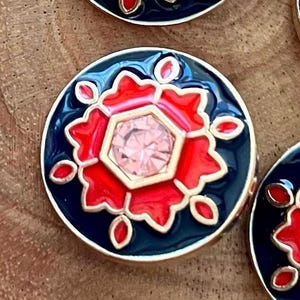 May include: A round, black button with a gold rim. The button has a red and gold design with a pink rhinestone in the center.