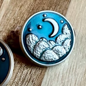 May include: A silver pin with a blue enamel background featuring a crescent moon and clouds. The clouds are textured and the moon is white.