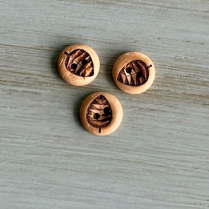 May include: Three round, light brown wooden buttons with leaf-like designs carved into their surfaces. Each button has two small holes for sewing. The buttons are arranged on a light gray surface.