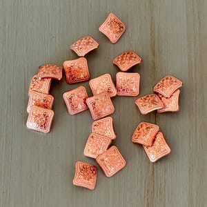 May include: A collection of copper-colored square and diamond-shaped metal beads with intricate embossed patterns. The beads are scattered on a light brown wooden surface, showcasing their detailed designs and warm metallic hue.