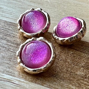May include: Three pink glitter buttons with gold metal rims. The buttons have a slightly raised, rounded shape.