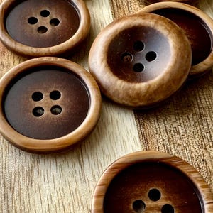May include: Close-up of several large, round buttons with four holes each. The buttons have a dark brown center and a lighter brown, wood-grain-like rim. The buttons are arranged on a wooden surface.