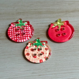 May include: Three decorative strawberry-shaped buttons. One button features a red and white gingham pattern, another is solid red, and the third has a pattern of small strawberries. Each button has a green stem and four holes for sewing.