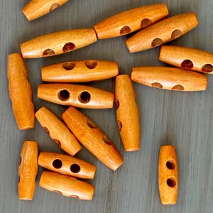 May include: A collection of light brown wooden toggle buttons with two or three holes each. The buttons are oval-shaped and have a smooth, polished finish. They are scattered on a gray surface.