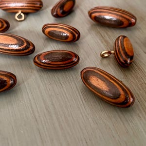 May include: A collection of 10 oval-shaped wooden buttons with brown and orange stripes. The buttons have a smooth, polished finish and are perfect for adding a touch of rustic charm to any project.