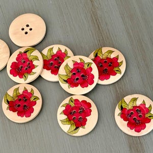 May include: A collection of wooden buttons with a floral design. The buttons are round and feature a light wood color. Each button has a red flower design with green leaves. One button has four holes for sewing.