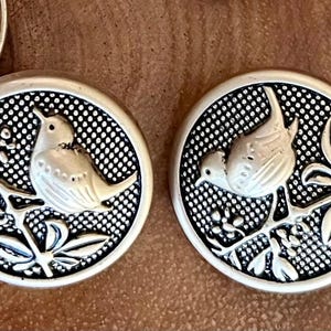 May include: Silver-toned metal buttons with a bird and foliage design. The buttons have a textured background and a raised relief of a bird perched on a branch with leaves and berries. The buttons are round.