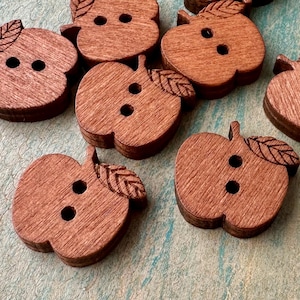 May include: Wooden apple-shaped buttons with two holes and a leaf detail. The buttons are a warm brown color and have a wood grain texture. These decorative buttons are ideal for sewing and craft projects.