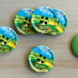 May include: A collection of six round buttons. Five buttons feature a landscape design with blue skies, green fields, and yellow accents. The sixth button is solid green with four holes.
