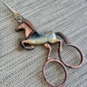May include: A pair of copper-toned metal scissors shaped like a horse. The horse's mane forms the blade, while the body features a blue and cream gradient finish. The two finger loops are integrated into the horse's hind legs and tail design.