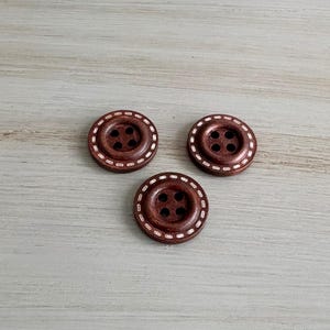 May include: Three brown wooden buttons with four holes each. The buttons have a decorative white dotted border around the edge. The buttons are arranged on a light wood-grain surface.
