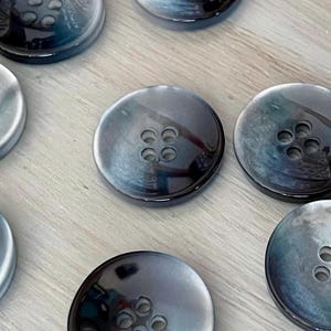 May include: A collection of round, four-hole buttons featuring a glossy, iridescent finish with swirling shades of dark blue, grey, and black. Each button has a smooth, polished surface and a centered square pattern of four small circular holes.