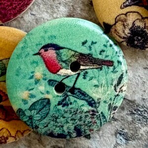 May include: A collection of round, flat buttons featuring printed illustrations of birds and floral patterns. The buttons are colored in shades of teal, yellow, and green, each with two central holes for attachment. The surfaces display detailed nature-themed graphics.