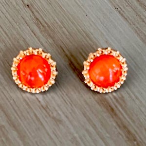 May include: A pair of orange stud earrings with a gold-toned metal setting. The earrings have a round, slightly raised design.
