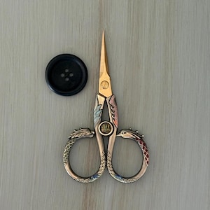 May include: A pair of gold-toned metal embroidery scissors featuring ornate dragon and phoenix handle designs with a central circular emblem containing Chinese characters, accompanied by a single round black four-hole sewing button.