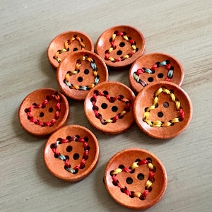 May include: A collection of wooden buttons with a heart-shaped design. Each button is a warm brown color and features a heart embroidered with colorful thread. The buttons have two holes for sewing and are arranged on a light gray surface.