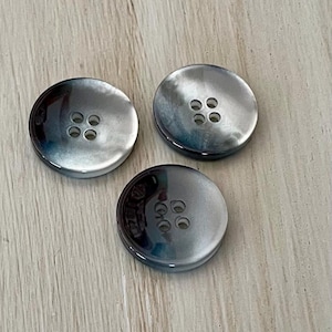May include: Three circular buttons with a smooth, polished finish. Each button features a four-hole design in the center and displays a gradient of dark charcoal gray to light silver tones with a subtle, pearlescent sheen across the surface.