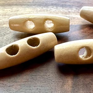 May include: Four natural wood toggle buttons with two holes each. The buttons are oval shaped and have a light brown color.