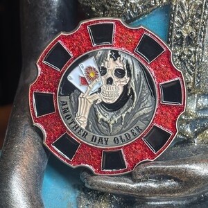 May include: A red and black poker chip with a skull wearing a hooded robe holding a playing card. The chip has the text "Another Day Older" around the edge.