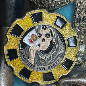 May include: A gold-colored metal poker chip with a black and gold glitter design. The chip features a black and white illustration of a skeleton holding two playing cards. The text "Another Day Older" is written around the edge of the chip.