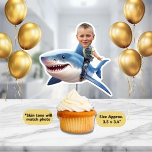 May include: A cupcake with white frosting topped with a shark-themed cake topper. The topper features a boy riding a blue shark. Gold balloons are in the background. Text on the image reads "Skin tone will match photo" and "Size Approx. 3.5 x 3.4".