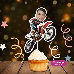 May include: A cupcake topped with white frosting, with a decorative topper featuring a boy riding a red and black dirt bike. The topper is approximately 4 x 2.8 inches. The background has bokeh lights and star and swirl decorations.