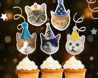 May include: Five cupcakes decorated with cat faces wearing party hats. The cats are wearing a variety of hats, including a crown, a blue and white striped hat, a rainbow hat, and a gold hat. The cupcakes are all white with white frosting.
