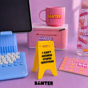 May include: A yellow miniature caution sign with the black text I CAN'T ANSWER STUPID QUESTIONS, positioned near a light blue tablet stand with a keyboard, a pink mug labeled BANTER, a pink printer, a clear water bottle, and a pink smiley face card.