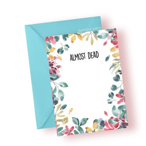 May include: A greeting card with the text "ALMOST DEAD" in black, surrounded by watercolor floral illustrations in pink, red, yellow, and green. The card is paired with a light blue envelope. The brand name "BANTERCARDS" is visible on the envelope.