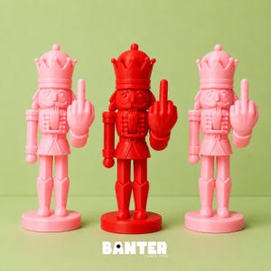 May include: Three nutcracker figurines standing in a row, with two light pink figures flanking a central bright red figure. Each nutcracker wears a crown and holds up a hand with the middle finger extended. The text BANTER CARDS.COM is visible.