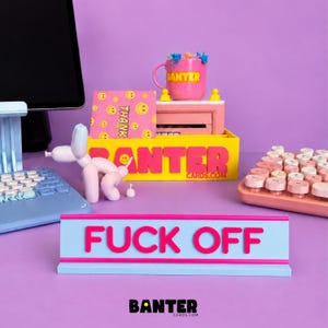May include: A light blue desk sign with the text FUCK OFF in bold pink letters. Behind it sits a yellow box labelled BANTER CARDS.COM, a pink mug with the word BANTER, a pink card with yellow smiley faces, and a light pink balloon dog figurine.