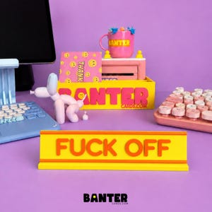 May include: A yellow desk sign with orange text reading FUCK OFF, positioned in front of a yellow organiser box labelled BANTER CARDS.COM. A pink mug with the word BANTER, a pink smiley face card, a light blue keyboard, and a pink keyboard are visible.