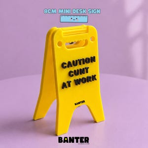 Puede incluir: Un letrero de escritorio en miniatura de color amarillo brillante en forma de A que mide 8 cm de altura. El letrero presenta letras negras en relieve que dicen CAUTION CUNT AT WORK. La palabra BANTER está impresa en letras negras pequeñas.