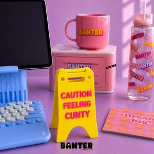May include: A light blue tablet stand with a keyboard, a pink mug with the word BANTER, a pink storage box, a yellow caution sign with the text CAUTION FEELING CUNTY, a clear water bottle with the word BANTER, and a pink card with the text Dream big.