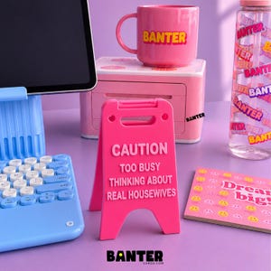 May include: A bright pink miniature caution sign with the text CAUTION TOO BUSY THINKING ABOUT REAL HOUSEWIVES, positioned next to a light blue tablet stand, a pink mug labeled BANTER, a pink printer, a water bottle, and a notepad with smiley faces.