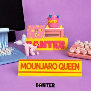 May include: A yellow desk nameplate with the orange text MOUNJARO QUEEN, a yellow organiser with pink text BANTER CARDS.COM, a pink mug with yellow text BANTER, a pink smiley face card, a light blue keyboard, a pink keyboard, and a balloon dog figure.